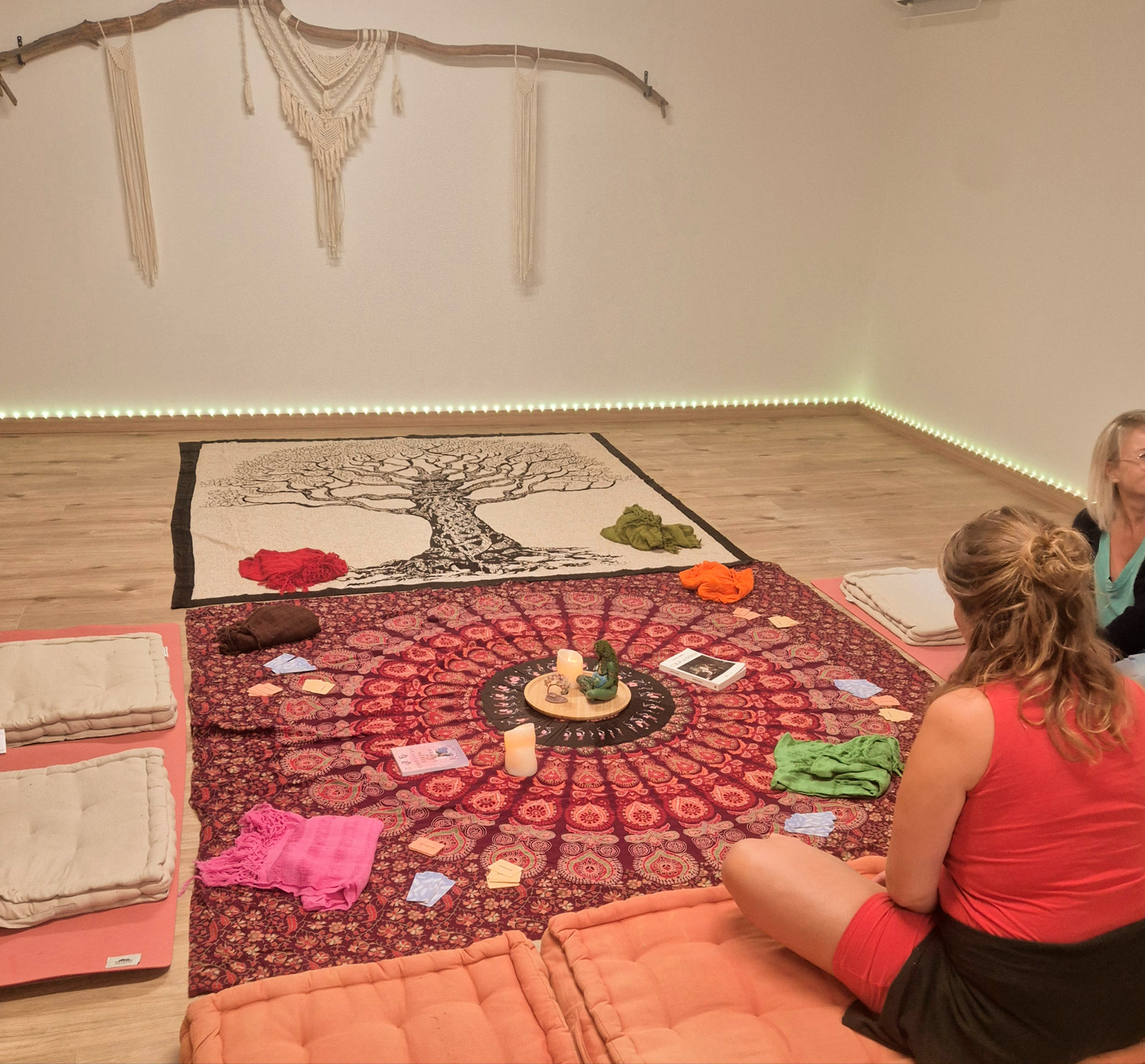 Salle d'atelier : tissu imprimé d'un arbre de vie au sol, cercle de mandala, châles rebozo pliés par couleur, coussins de sol ; deux femmes assises