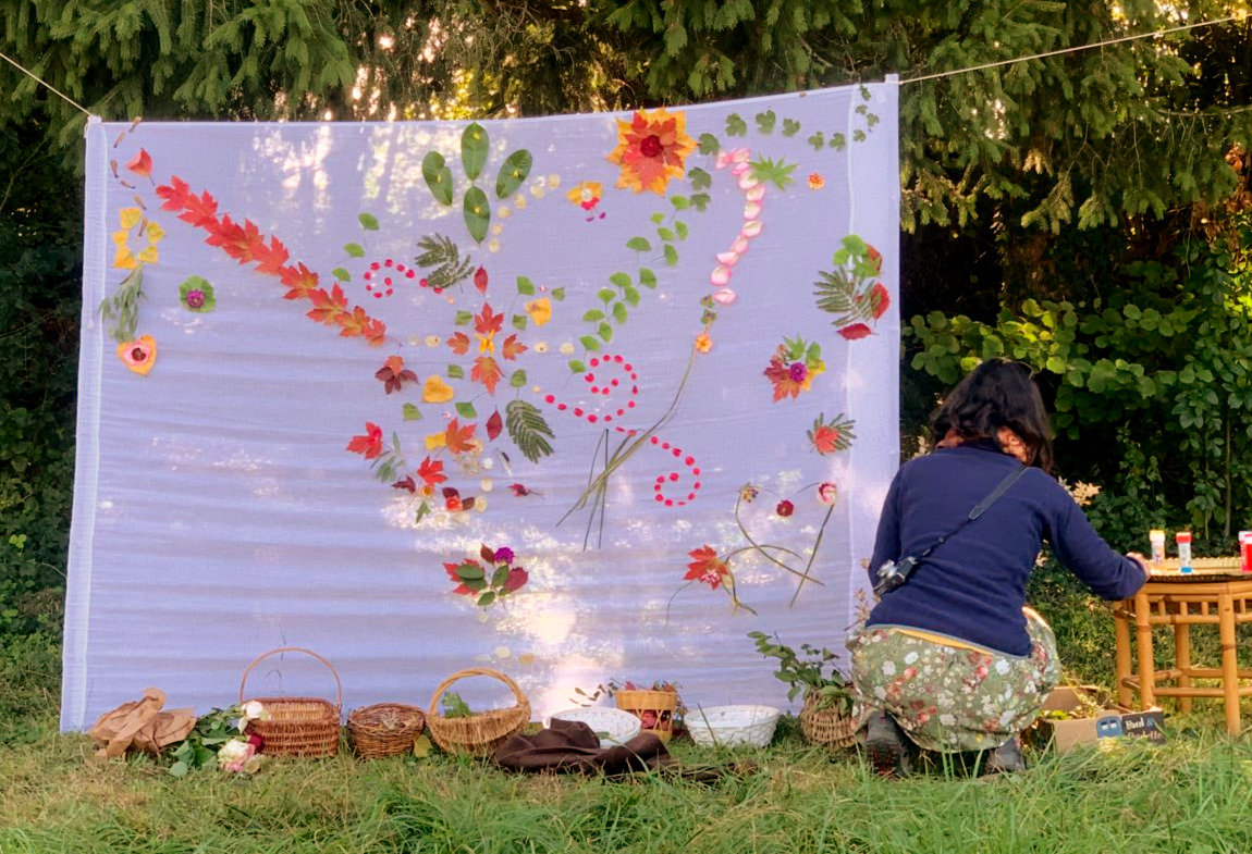 Grande toile claire tendue entre des arbres, couverte d'une composition de feuilles, fleurs et pétales en forme d'oiseau ; une femme accroupie prépare les matériaux, des paniers d'osier au sol