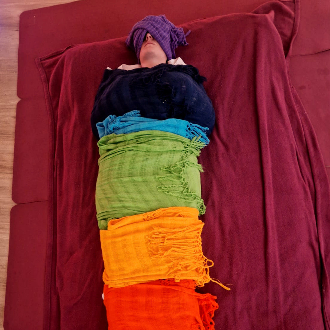 Personne allongée sur un matelas, enveloppée de la tête aux pieds dans une succession de châles rebozo violet, bleu marine, turquoise, vert, orange et rouge