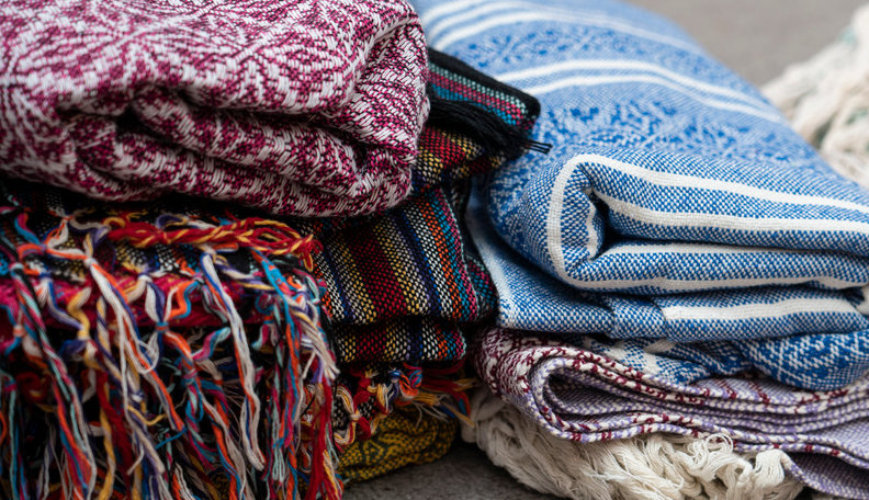 Pile de châles rebozo pliés : chevrons bordeaux et blancs, rayures multicolores à franges, tissage bleu et blanc