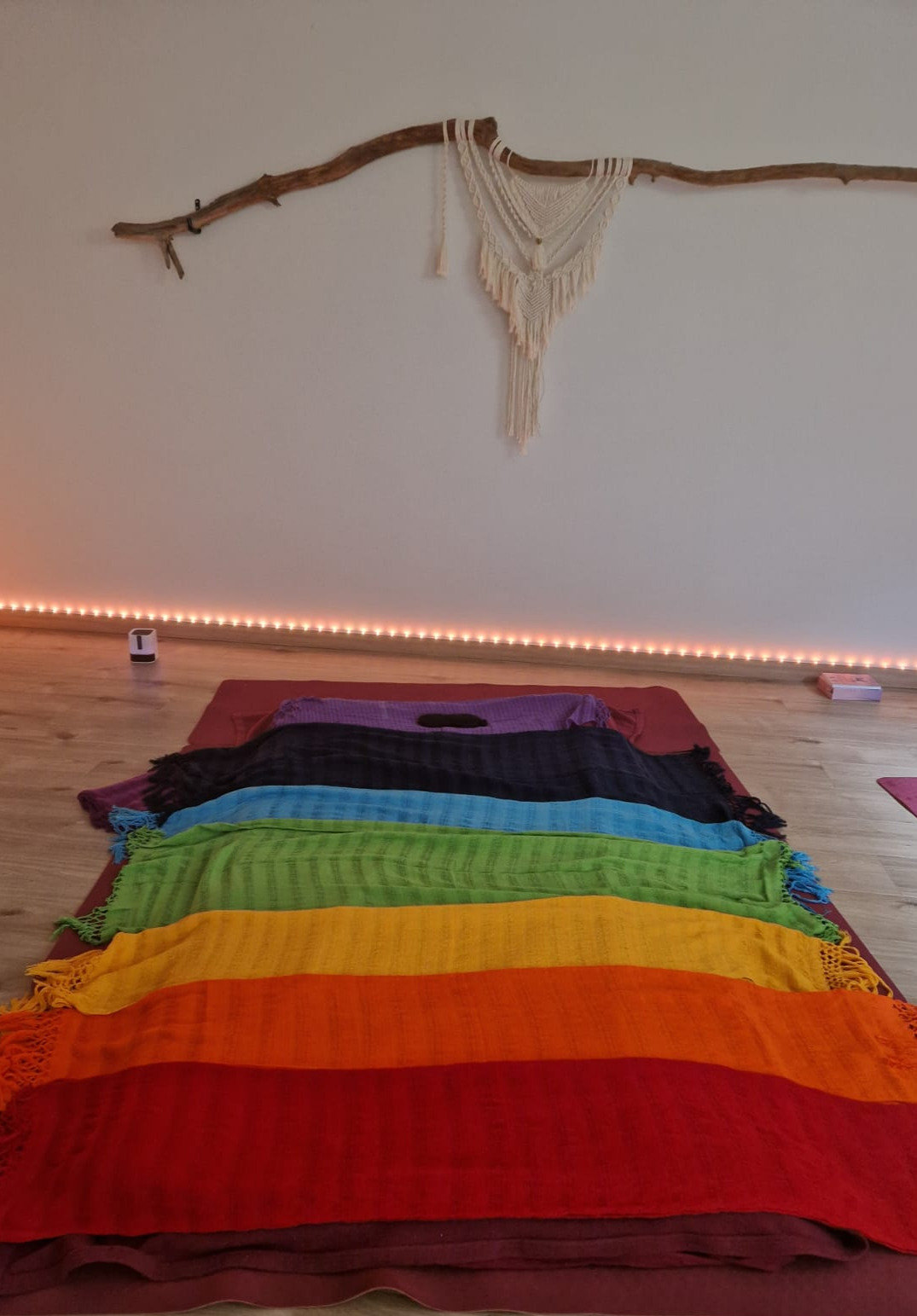 Salle de soin préparée : un matelas au sol recouvert de châles rebozo disposés en dégradé de couleurs, une suspension en macramé sur une branche de bois flotté
