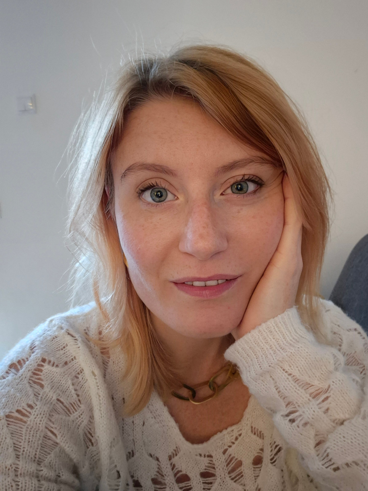 Portrait de Manon, souriante, la main contre la joue, devant un mur clair