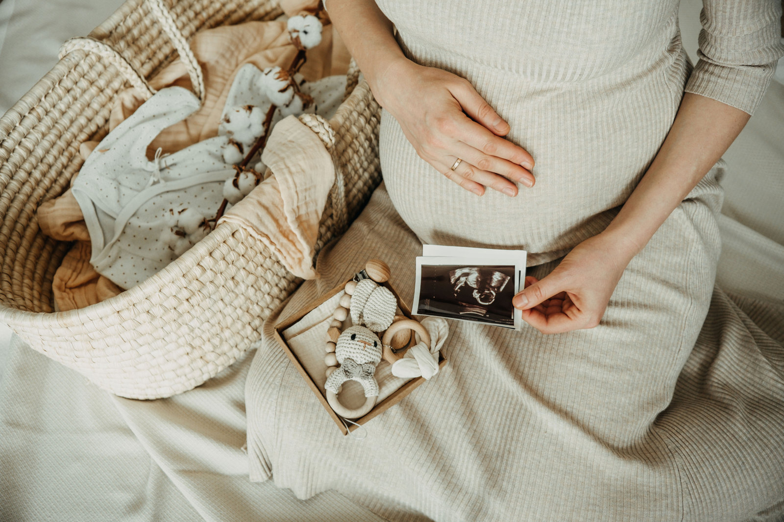 Vue de dessus : une femme enceinte, une main posée sur son ventre, tient une échographie ; à côté, un couffin en osier garni de linge, une branche de coton et un coffret de jouets en crochet et en bois.